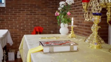 Altın sunak masası, kutsal kitapla süslenmiş şamdanlar ve sıcak tuğla duvara dayanan tek bir mum, güllerle işlenmiş beyaz vazo, işlemeli kumaş ve parlatılmış pirinç kutsal ayinsel atmosfer yaratıyor.