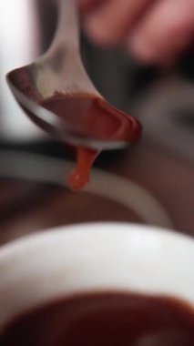 Yakın çekim kaşığı bardağa çikolata serpiştiriyor, kepçeden ipeksi kurdele düşüyor, sıcak kakao yüzeyi, zanaatkar baristalar dolduruyor, rahat mutfak ışıklandırması, yavaş duyusal hareket ve parlak doku.