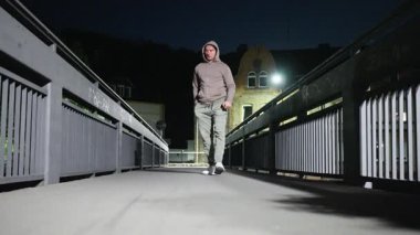 Gece vakti köprüde dikilen kapüşonlu bir adam kırmızı bardağı sokak lambasının altına atar.,