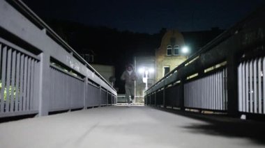 Adam gece köprüde koşuyor, alçak açılı yürüyüş yolu metal parmaklıklar ve uzak aydınlatılmış evle vurulmuş.,