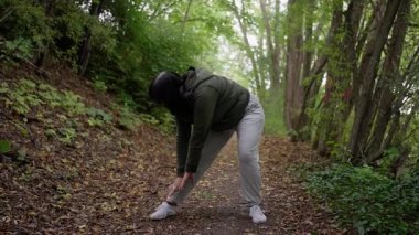 Kafkas kadın ayakkabıyı ağaçlık yolda bağlamak için eğiliyor, odaklanmış bağcıklar ve ısınma hareketleri, çömelme ve gerilme sekansı, koşucu hazırlık ve zindelik rutini, toprak yapraklar ve antrenman hazırlığı