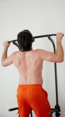 Üstsüz Asyalı adam parlak ev stüdyosunda barfiks çekiyor, turuncu şortlar, odaklanmış sırt kasları, barfiks çekme, kontrollü tekrarlar, ter ve düzenli nefes alma, atlet eğitim rutini,