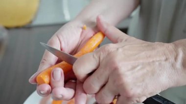 Beyaz tabağın üzerinde havuç soyan beyaz eller, keskin bıçak darbeleriyle derisi yüzülmüş parlak turuncu kökler, sebze artıkları ve sıcak gün ışığında günlük mutfak sahnesi, taze malzemeler hazırlamak.