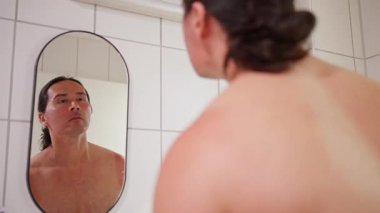 Üstsüz beyaz adam fayanslı banyo aynasında yansımayı kontrol eder, ıslak saç, tıraş losyonu sürme, esnek kol, cilt değerlendirmesi, kendine güvenen ifade, samimi sabah bakım ritüeli, yumuşak doğal