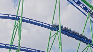 Hız trenine binenler mavi pist yeşil çelik desteğini artırıyor, arabalar heyecanlı yolcularla dolu, eller havada, neşeli çığlıklar, yüksek irtifa eğrisi, parlak yaz gökyüzü, sinematik çerçeve. 4k