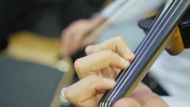 Prova sırasında keman çalan yakın plan eller, odaklanmış parmak izleri ve arka planda bulanık müzisyenlerle bir konser salonu provasında baş eğmek, sıcak ahşap enstrüman, ayrıntılı mandal kutusu.