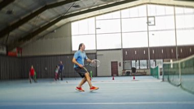 Adam kapalı sahada agresif bir şekilde hizmet veriyor, havada hareketlilik var, güçlü bir salıncak ve takip et, spor ayakkabıları yüzeyi kazıyor, koçun gözlemlediği yoğun eğitim tatbikatı.