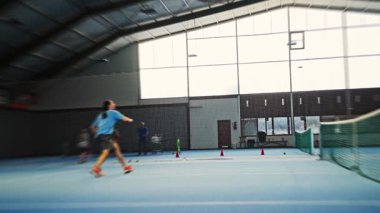 Oyuncu mavi sahada koşuyor, keskin yan ayak egzersizleri ve reaktif antrenmanlar, koç hızlılık için topları atıyor, yüksek tempolu çeviklik odağı, toz yutan spor ayakkabıları