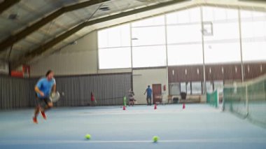 Adam kapalı sahada topları topluyor, beslenme talimi yapıyor, raketi ayarlıyor, tavan penceresinin altında alçak çekimler için eğiliyor, taban çizgisinde koniler, tek başına ısınma sırasında kararlı atmosfere odaklanıyor.