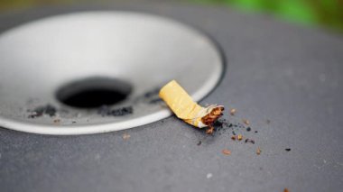 discarded cigarette butt on ashtray metal surface, closeup of filter end and ash crumbs beside circular disposal hole, urban park background blurred, mood of neglect and small-scale pollution, muted