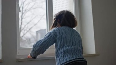 Genç bir çocuk pencereden dışarı bakıyor, yansımayı teşvik eden sakin bir ortamda merak ve tefekkür uyandırıyor. Yavaş çekim