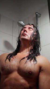 Duş başlığının altındaki adam ıslak kaslar, gövdesi parlayan, başı öne eğilmiş, gözleri kapalı su yüzüne ve göğsüne hücum ederken, tuvalette buhar yükseliyor, derin düşüncelere dalmış, mahrem bir ifade.