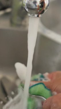 Ev Mutfağındaki Musluğun Altında El Yıkama Süngeri, Paslanmaz Musluktan akan ılık su Yeşil Süngerin üstüne köpüklü sabun köpüğü Parmaklarına köpüklü sabun köpüğü, Havzada dinlenen kaşık,