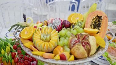 Papaya, üzüm ve nar içeren canlı meyve tabağı, düğün büfesinde gümüş tepsiye işlenmiş oyulmuş kavun ve mango dilimleri, yumuşak doğal ışık, zarif sofra takımı ve şenlikli atmosfer