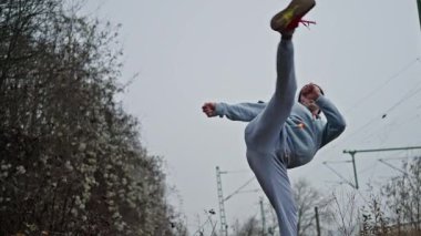 Beyaz kadın açık havada yüksek tekme atıyor zarif ama etkili teknik, kapüşonlu ve eşofman altı, süpürme dairesi ve tam ekran, sabit bakış, demiryolu kablolarıyla kaplanmış gökyüzü,