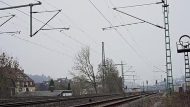 Bulutlu Demiryolu Rayları Köye yakın, Boş Raylar Catenary Polonyalıların altında Yapraksız Ağaçlar, Rutubetli Denge ve Çakıllar, Uzak Evler ve Endüstriyel Sinyaller, Sessiz Kış Paleti,