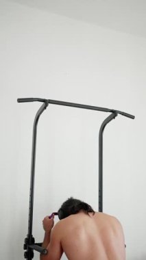 Gömleksiz Adam Eğitimi Çekme Çubuğu Arka Görünümü, Turuncu Şort ve Kulaklıklar Tutuşunu Ayarlıyor ve Kontrollü Barfiks Tekrarı, Beyaz Duvara Karşı Güçlü Sırt Kasları Kasılmaları, Minimalist