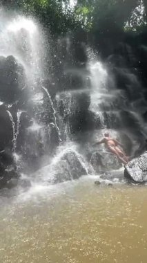 Şelale gibi akan tropik şelalenin altında dikilen, püskürten ve sisli Rocky Ledge, güneşli gölgeli gezgin poz veren, enerjik su akıntısı, yağmur ormanı arkaplanı ve hayat dolu