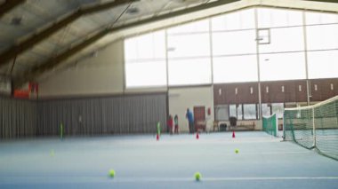 Kapalı Tenis Sahası Oyuncuları Antrenman Konileri ve Balo Tatbikatları, Koç Acemi Adaylara Ağ Yakınları, Büyük Pencerelerden Gün Işığı, Dağılmış Topları ve Isınma Esnemeleri Olan Mavi Hardcourt