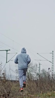 Açık otlak arazide koşan sporcu giysisi. Kapüşonlu ve Koşu manevrası yapan sporcu figürü ıssız kırsal bölgelerde uzak demiryolu yapıları ile manevra yapıyor.