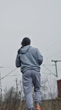 Koşu Kadın Eğitimi. Atletik Kadın Parmaklıklar Arasında Koşuyor. Kapüşonlu beyaz kadın koşucu adımlarına odaklanıyor. Antrenman sırasında tren yolunun yanında sabit bir adım atarken at kuyruğu sallanıyor.