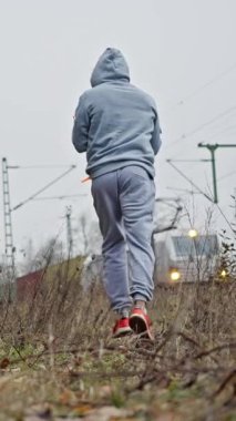 Training along Tracks 'da. Adam Sabit Sanayi Arkaplanı 'nı işletiyor. Tren yolu boyunca koşan kapüşonlu bir erkek atlet. Demiryolunda Başlıklı Disiplinli Erkek Koşucu Dayanıklılık Göstergesi