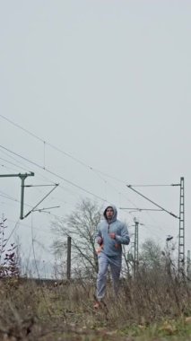 Dışarıdaki Adam Eğitimi Demiryolu yakınında Koşu, Greyhoodie Koşucusu Soğuk Alan 'da Güç Kullanımı, Görünür Nefes, Sert Çimenler Üzerinde Sabit Adımlar, Uzak Sütunlar ve Kablolar, Gritty Bulut Enerjisi, Odaklanmış Enerji