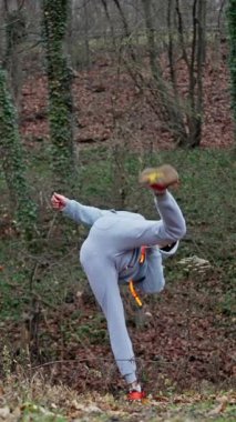 Beyaz Adam Patlayıcı Atlama Tekmesi Eğitimi. Havadan Dönüş ve Odaklı İniş, Hızlı Toparlanma Basamağı, Kapüşonlu ve Gri Eşofman, Soğuk Orman Zemininde Kırmızı Koşu Ayakkabıları, Solo Fighter