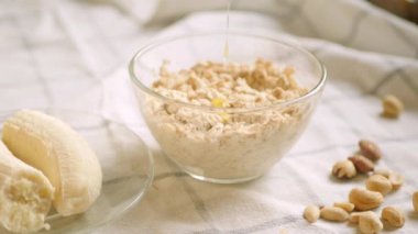 Bir gecelik yulaf ezmesi kasesi. Evde sağlıklı kahvaltı hazırlıyorum. Bitki sütü dökülüyor. Muz, bal ve fındık malzemeleri. 4K video. Adım adım tarif