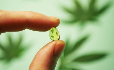 Close up hand holding CBD oil pill. Cannabis leaves reflection. Medical marijuana usage  clipart