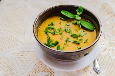 Dantelli arka planda yeşil ıspanak ve nane ile süslenmiş zengin, kalın balkabağı çorbası dolu kahverengi çizgili bir kase oturuyor..