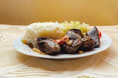 Beyaz pirinç, dilimlenmiş domates, doğranmış lahana ve sığır ciğeri kahvaltısı soluk sarı bir duvara karşı bağlanmış masa örtüsüyle kaplı küçük beyaz bir çiçek tabağında servis edilir...