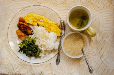 Kızarmış muzdan yapılmış bir kahvaltı, kızarmış yumurta rulosu, buharda pişmiş amaranth ve şeffaf kırılabilir bir yerde, dantelli masa örtüsüyle kaplı bir masada haşlanmış beyaz pirinç..