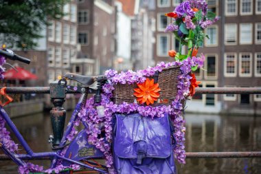 30 Haziran 2021, Amsterdam, Hollanda, Kanal köprüsüne parkedilmiş renkli yapay çiçeklerle süslenmiş bisiklet, geleneksel mimari evlerin arka planında bulanıklık