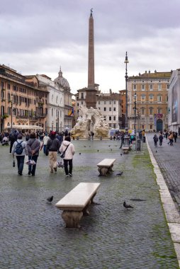19 Ocak 2026, Roma, İtalya - Roma 'da bulutlu kış gökyüzünün altında güvercinler kaldırım taşlarının üzerinde toplanırken, insanlar Dört Nehir Çeşmesi ve dikilitaşın yanındaki Piazza Navona Meydanı' nda yürür.