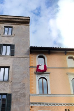 18 Ocak 2026, Roma, İtalya: Kırmızı ve beyaz yatak, parçalı bulutlu bir kış gökyüzüne karşı hazırlanan pastel cepheler arasındaki pencere pervazında asılı.