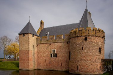 26 Mart 2023 - Medemblik, Hollanda - Radboud Müzesi, yuvarlak kulesi ve kayrak çatıları olan tarihi tuğla kale, bulutlu bir yay altında sakin bir hendeğin yanında duruyor.