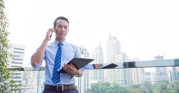 Smart Asian Business man using smart phone and document to working on window with city building and green balcony