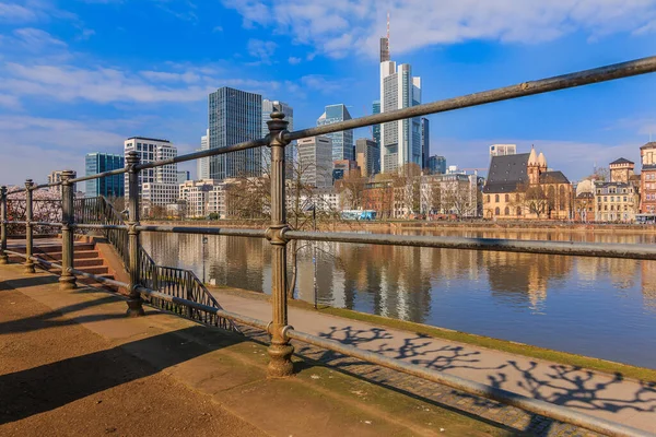 Financial center with skyscraper of Frankfurt am Main. River with railings in the foreground on a sunny day. Bank of the river with reflections in spring with blue sky