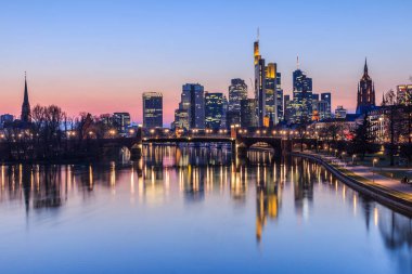Akşamları Frankfurt silueti. Mavi saatte gün batımında finans ve iş bölgesinden aydınlık gökdelenlerle. Nehirdeki yansımalar Kıyıdaki parkla