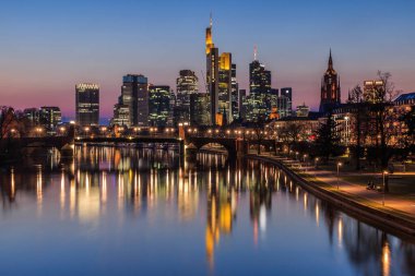Frankfurt silueti akşam mavi saatte. River Main 'de yüksek binaların yansımaları var. Nehir kıyısında bir köprü ve parkla aydınlatılmış ticari binalar.