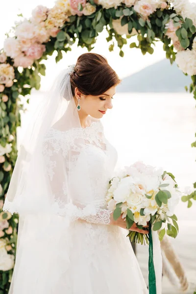 Bella sposa con un velo e bouquet da sposa vicino all'arco di nozze — Foto Stock