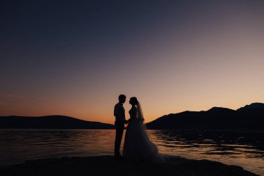 Gelin ve damat gün batımında suyun yanındaki iskelede el ele tutuşuyorlar.