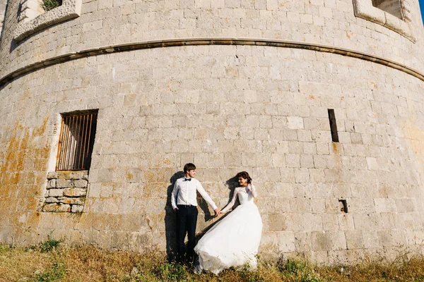 Pengantin wanita dan pengantin pria berpegangan tangan dan berdiri di dekat dinding benteng Arza — Stok Foto