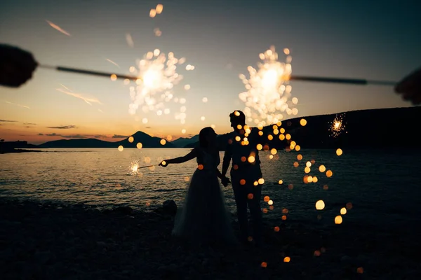 La mariée et le marié debout sur la plage de l'île de Mamula et tenant des étincelles au coucher du soleil — Photo