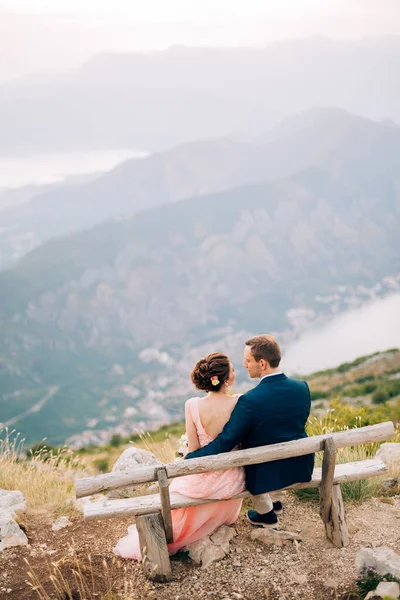 Novia y novio sentado en el banco en la montaña contra el telón de fondo de la bahía de Kotor —  Fotos de Stock