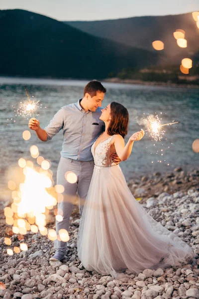 La mariée et le marié s'embrassant sur la plage de l'île de Mamula et tenant des étincelles au coucher du soleil — Photo
