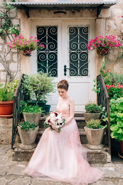 Pengantin cantik dalam gaun pengantin lembut dengan buket pengantin duduk di teras rumah di Perast — Stok Foto