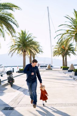 Baba ve kızı deniz kıyısındaki palmiye ağaçlarının yanında el ele koşuyorlar.