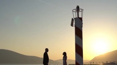 Gelin ve damat Porto Karadağ 'da deniz fenerinin yanındaki iskelede kucaklaşıyorlar. Üzerlerinde bir uçak uçuyor.